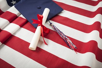 Graduation cap with tassel and red ribbon wrapped diploma resting on american flag