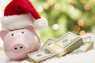 Pink piggy bank wearing santa hat near stacks of hundreds of dollars of money on snowflakes