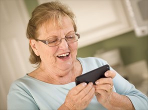 Happy senior adult woman texting on her smart cell phone in kitchen
