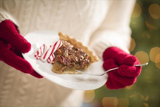 Woman wearing A sweater and seasonal red mittens holding A plate of pecan pie with peppermint sticks