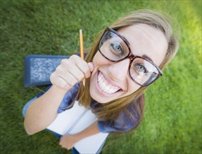 Fun wide angle portrait of pretty young woman with books and pencil sitting in the grass outdoors