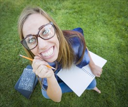 Fun wide angle portrait of pretty young woman with books and pencil sitting in the grass outdoors