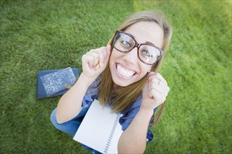 Fun wide angle portrait of pretty young woman with books and pencil sitting in the grass outdoors