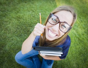 Fun wide angle portrait of pretty young woman with books and pencil sitting in the grass outdoors