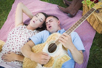 Happy mixed-race couple on picnic blanket at the park playing guitar and singing songs