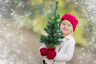 Baby girl in red mittens and cap holding small christmas tree outdoors with snow effect