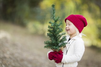 Baby girl in red mittens and cap holding small christmas tree outdoors