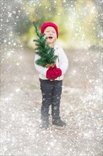 Baby girl in red mittens and cap holding small christmas tree outdoors with snow effect