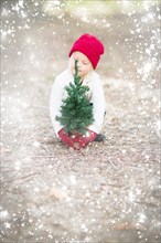 Baby girl in red mittens and cap near small christmas tree outdoors with snow effect