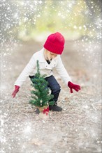 Baby girl in red mittens and cap near small christmas tree outdoors with snow effect