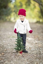 Baby girl in red mittens and cap near small christmas tree outdoors