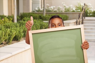 Happy hispanic boy with thumbs up holding blank chalk board outside on school campus