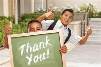 Happy hispanic boys giving thumbs up holding thank you chalk board outside on school campus