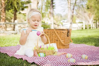 Cute baby girl enjoys coloring her easter eggs on picnic blanket in the grass