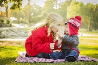 Little girl with her baby brother wearing winter coats and hats sharing a lollipop outdoors at the park