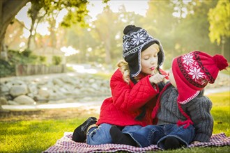 Little girl with her baby brother wearing winter coats and hats sharing a lollipop outdoors at the park