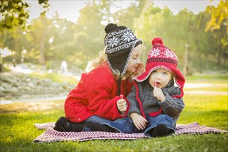 Little girl whispers A secret to her baby brother wearing winter coats and hats sitting outdoors at the park