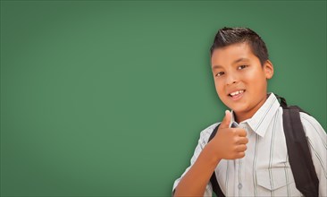 Cute hispanic boy with thumbs up wearing backpack in front of blank chalk board
