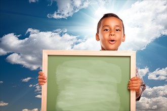 Proud hispanic boy holding blank chalkboard over blue sky and clouds with sun burst