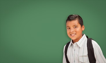 Cute hispanic boy with backpack in front of blank chalk board
