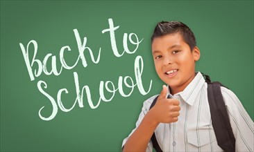Cute hispanic boy with thumbs up wearing A backpack in front of chalk board with back to school written on it