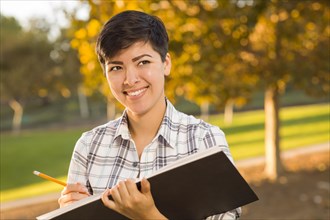 Portrait of a pretty mixed-race female holding sketch book and pencil outdoors at the park on a sunny afternoon