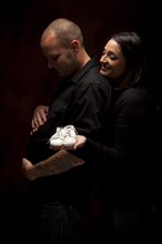Fun mixed-race couple holding new white baby shoes against a black background under dramatic lighting