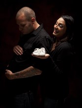 Fun mixed-race couple holding new white baby shoes against a black background under dramatic lighting