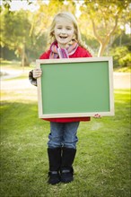 Cute little blonde wearing winter coat and scarf holding a green chalkboard outdoors