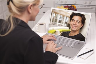 Happy young woman in kitchen using laptop online dating search with portrait of man on screen
