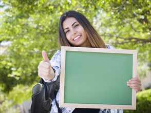 Portrait of an attractive excited mixed-race female student holding blank chalkboard and carrying backpack on school campus