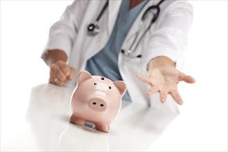 Demanding doctor wearing stethoscope reaches palm out behind piggy bank