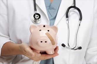 Female doctor with stethoscope holding piggy bank with bandage on face