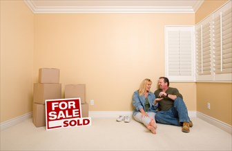 Excited couple with new keys relaxing on floor near boxes and sold real estate signs in empty room