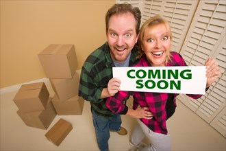 Goofy couple holding coming soon sign in room with packed cardboard boxes