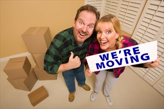 Goofy thumbs up couple holding we're moving sign in room with packed cardboard boxes