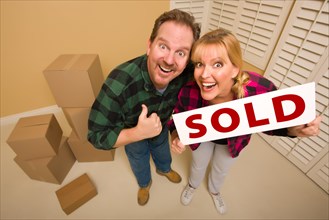 Goofy thumbs up couple holding sold sign in room with packed cardboard boxes