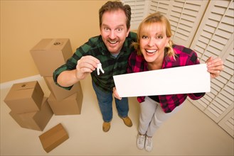 Goofy couple holding keys and blank sign in room with packed cardboard boxes