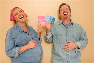 Laughing man and pregnant woman deciding on pink or blue wall paint with swatches in hand