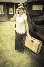 Happy 1920s dressed girl holding suitcase next to vintage car and cabin