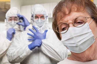 Senior adult woman wearing medical face mask in doctor office with nurses in HAZMAT suits behind