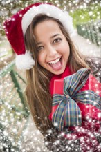 Pretty festive smiling woman wearing a christmas santa hat with bow wrapped gift outside with snow flakes border