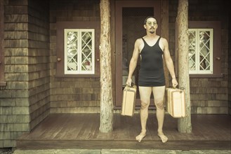 Goofy gentleman dressed in 1920's era swimsuit holding suitcases on porch of cabin
