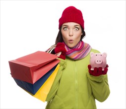 Concerned expressive mixed-race woman wearing winter clothes holding shopping bags and piggybank isolated on white background
