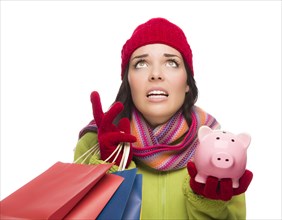 Stressed mixed-race woman wearing winter clothing looking up holding shopping bags and piggybank isolated on white background