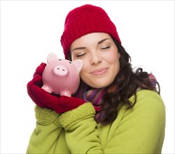 Pleased mixed-race woman wearing winter clothing hugging piggybank isolated on white background