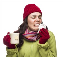 Sick mixed-race woman wearing winter hat and gloves blowing her sore nose and holding cup of hot tea isolated on white