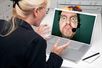 Shocked woman in kitchen using laptop with grumpy customer support man on screen