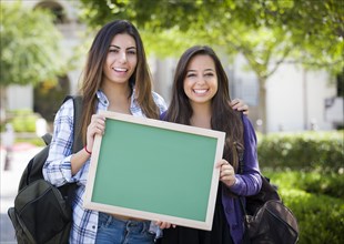 Portrait of two attractive mixed race female students holding blank chalkboard and carrying backpacks on school campus