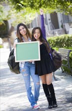 Portrait of two attractive excited mixed race female students holding blank chalkboard and carrying backpacks on school campus